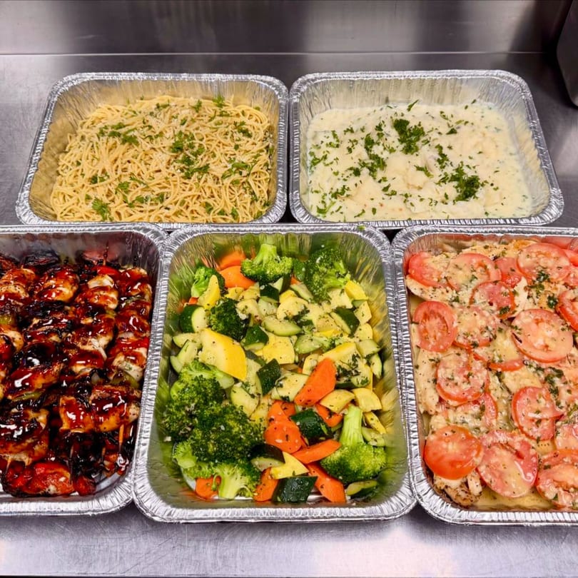 Five aluminum trays of food, including spaghetti, mashed potatoes, chicken skewers, mixed vegetables, and chicken with tomatoes.
